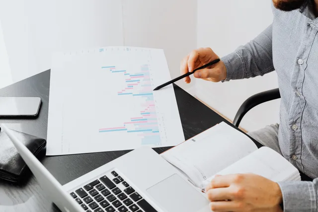 An office setup with a person analyzing a graph printout while using a laptop and notebook. Barometr zawodów: prognozy a statystyka rynku pracy